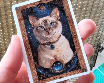 May include: A white cat with blue eyes is depicted on a card with a gold frame. The cat is wearing a necklace with a blue gemstone. The background is a dark blue with gold stars and a crescent moon.