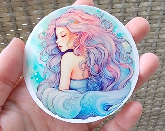 May include: A round sticker featuring a watercolor illustration of a mermaid with long pink and purple hair. The mermaid is depicted with her eyes closed and her tail is curled around her body. The background is a light blue with white swirls and dots.