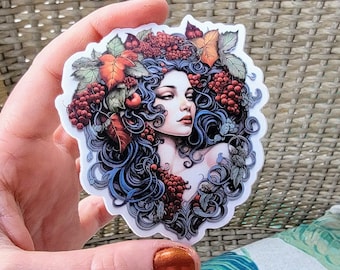 Op de afbeelding: Een sticker met een vrouw met lang blauw haar, omringd door rode bessen, groene bladeren en herfstbladeren. De vrouw heeft een zachte, dromerige uitdrukking en kijkt naar rechts.