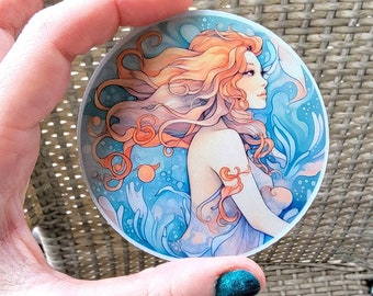 May include: A round sticker featuring a watercolor illustration of a woman with long, flowing red hair. She is surrounded by blue and white water-like designs.