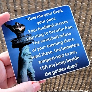 May include: A blue square coaster featuring an image of the Statue of Liberty's torch and the poem "The New Colossus." The text is in white and the image is set against a blue background. The coaster is held by a hand.