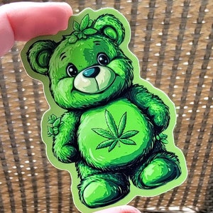 May include: Green plush teddy bear sticker with a marijuana leaf on its chest. The bear has a friendly expression and is wearing a flower crown.
