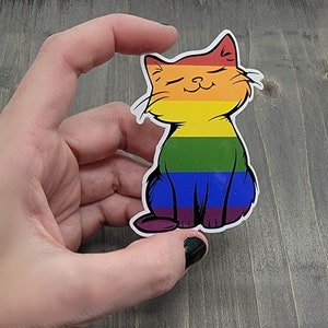 Op de afbeelding: Een regenboog-pride-katten sticker met een glimlachend gezicht. De kat zit met gekruiste poten en heeft een regenboogkleurig lichaam.
