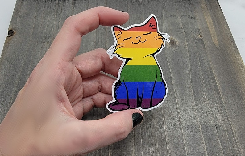 Op de afbeelding: Een regenboog-pride-katten sticker met een glimlachend gezicht. De kat zit met gekruiste poten en heeft een regenboogkleurig lichaam.