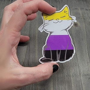 Op de afbeelding: Een sticker met een cartoonkat in een geel, wit, paars en zwart kleurenschema. De kat zit met zijn ogen dicht en een glimlach op zijn gezicht. De kleuren van de kat vertegenwoordigen de non-binaire pride-vlag.