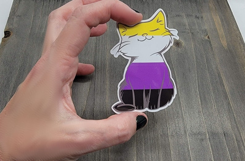 Op de afbeelding: Een sticker met een cartoonkat in een geel, wit, paars en zwart kleurenschema. De kat zit met zijn ogen dicht en een glimlach op zijn gezicht. De kleuren van de kat vertegenwoordigen de non-binaire pride-vlag.