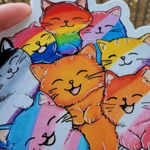 May include: A sticker featuring a group of cartoon cats with different colored stripes representing various pride flags. The cats are all smiling and have their eyes closed.