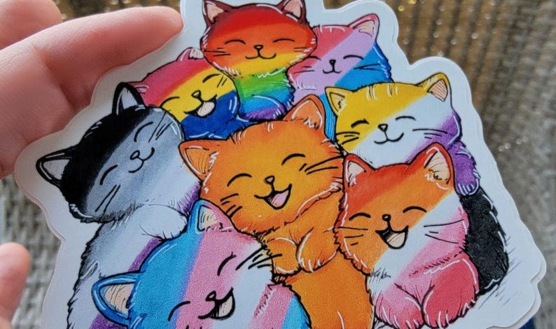 May include: A sticker featuring a group of cartoon cats with different colored stripes representing various pride flags. The cats are all smiling and have their eyes closed.