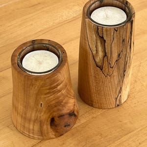Puede incluir: Dos candelabros de madera con un acabado natural de vetas. Cada soporte tiene un inserto de vidrio que contiene una vela blanca. Los candelabros tienen diferentes formas, uno es más redondeado y el otro más cilíndrico.