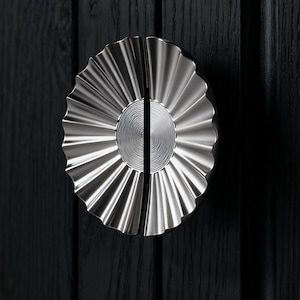 May include: A pair of silver cabinet handles with a fluted, sunburst design. The handles are mounted on a dark wood cabinet door. The handles are round with a central, circular design.