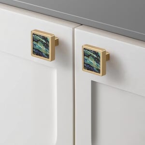 May include: Two square cabinet knobs with a gold finish and a colorful, iridescent inlay. The knobs are mounted on white cabinet doors. The inlay features shades of blue, green, and black, adding a decorative touch to the hardware.