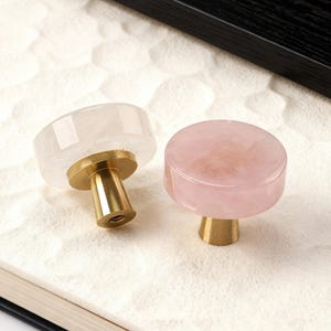 May include: Two decorative cabinet knobs. One is clear, the other is pink, both with a round shape and brass base. The knobs are made of a translucent material, possibly crystal or stone, and are set against a textured white surface.