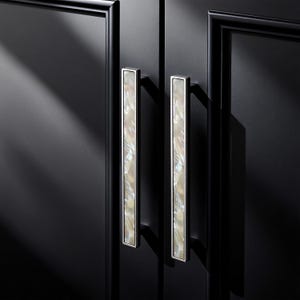 Puede incluir: Primer plano de dos tiradores de puerta de armario. Los tiradores son rectangulares con un marco plateado y un centro nacarado e iridiscente. Las puertas del armario son negras con un acabado brillante.