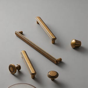 May include: An assortment of antique brass cabinet hardware, including handles and knobs. The hardware features a rectangular design with grooved detailing and a warm, aged finish. The collection includes various sizes and shapes for versatile use.