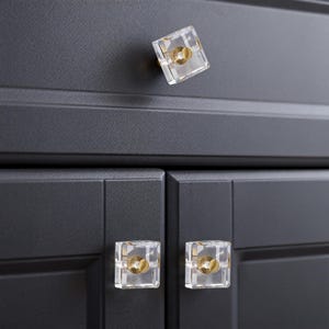 May include: Close-up of a black cabinet with three clear, square crystal knobs with gold-colored hardware. The knobs are evenly spaced on the cabinet doors and drawer, providing a modern and elegant aesthetic.