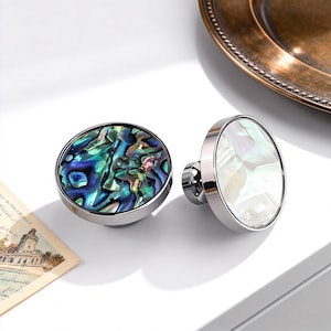 May include: Two decorative cabinet knobs. One features a vibrant, iridescent abalone shell design in shades of blue, green, and purple, set in a silver-toned metal. The other knob has a white, pearlescent surface, also set in silver. Both are oval-shaped.