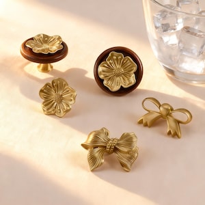 May include: A selection of decorative cabinet knobs and pulls in gold tones. The image shows flower and bow designs, some mounted on dark wood. These are suitable for furniture and home decor applications.