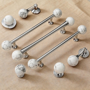 Puede incluir: Una colección de herrajes para armarios, que incluye tiradores largos y pomos redondos. Los herrajes presentan esferas de mármol blanco con vetas grises, combinadas con bases y barras de metal cromado o plateado pulido. Los herrajes se muestran sobre una superficie de tono neutro.
