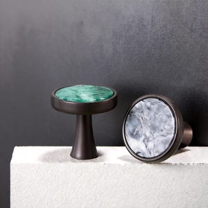 Könnte beinhalten: Zwei dekorative Schrankknöpfe. Einer hat eine grün marmorierte Oberfläche, der andere ein grau-weiß marmoriertes Muster. Beide Knöpfe haben eine dunkelbronze Oberfläche und sind vor einem dunklen Hintergrund platziert.