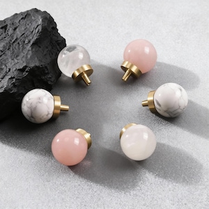 May include: A selection of decorative cabinet knobs with spherical tops in various colours and patterns. The knobs include clear, pink, white, and marble-like designs, each with a brass-coloured base and stem. These knobs are ideal for adding a touch of elegance to furniture.