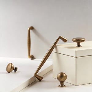 以下が含まれることがあります： アンティーク真鍮のキャビネット金具の品揃え。つまみと取っ手が含まれています。白い表面に配置され、背景には白い箱があります。真鍮はヴィンテージ感のある外観です。