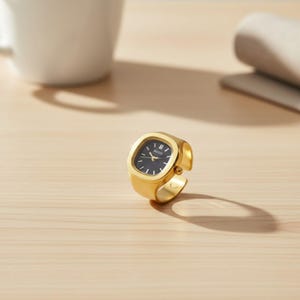 May include: A gold ring watch with a square black face. The watch features a minimalist design with a gold band. The watch face displays the time with gold hands and markers. This ring watch is a unique piece of jewellery. The watch is approximately 2 cm in length.