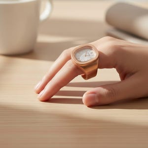 May include: A gold-coloured ring watch with a square face and white dial is worn on a finger. The watch has a minimalist design, with a simple face and hands. The ring is made of a shiny metal and appears to be a unique piece of jewellery.
