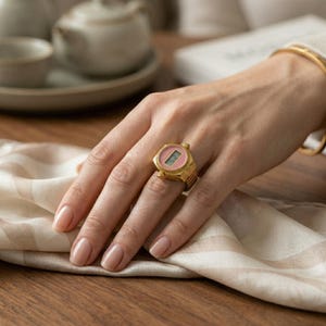 May include: A gold-coloured ring featuring a pink digital watch face. The ring has a rectangular shape with a ridged bezel and a digital display showing the time. The ring is on a hand with a gold bracelet. The background includes a tea set and a patterned cloth.