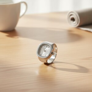 May include: A silver ring watch with a square face and a light-coloured dial. The watch has a band that wraps around the finger. The watch is on a light-coloured wooden surface.