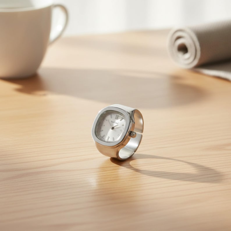 May include: A silver ring watch with a square face and a light-coloured dial. The watch has a band that wraps around the finger. The watch is on a light-coloured wooden surface.