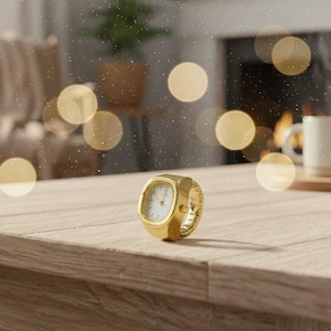 May include: A gold ring watch with a rectangular face and a white dial. The watch has a stretch band. It is displayed on a wooden surface. The background features blurred bokeh lights.