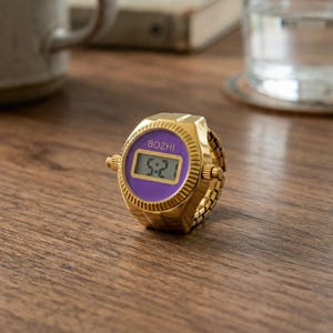 May include: A gold-colored digital ring watch with a purple face displaying the time 5:21. The watch has the brand name "BOZHI" above the time. The ring band is made of small, connected gold links.