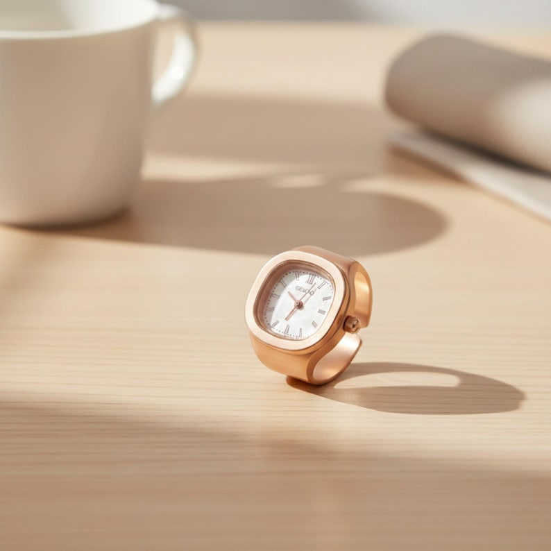 May include: A rose gold ring watch with a square face and white dial. The watch has a minimalist design and is set on a light wooden surface. A white mug and rolled paper are in the background.
