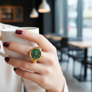 May include: A gold-toned ring watch with a green face and Roman numeral hour markers. The watch ring is being worn on a finger. The background is blurred, suggesting a cafe setting.