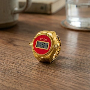 May include: A gold-colored ring watch with a red face displaying the time 5:21. The watch has the brand name "BOZHI" printed on the face. The ring band is made of small, connected gold links.