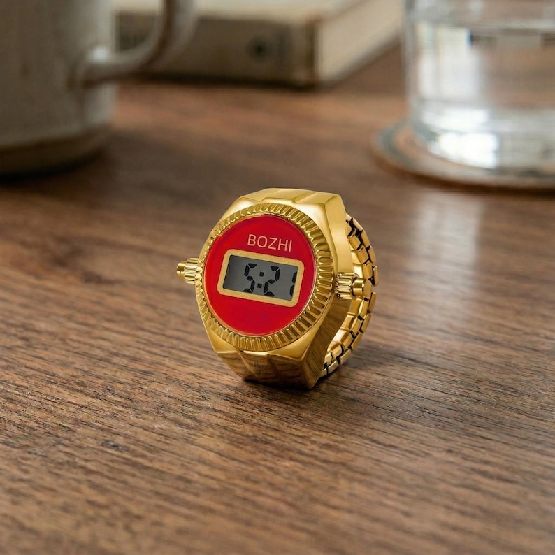 May include: A gold-colored ring watch with a red face displaying the time 5:21. The watch has the brand name "BOZHI" printed on the face. The ring band is made of small, connected gold links.