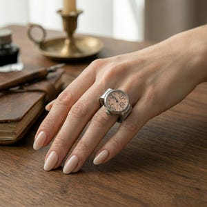 Könnte beinhalten: Eine silberfarbene Ringuhr mit hellrosa Zifferblatt und schwarzen Ziffern wird an einer Hand getragen. Die Uhr hat ein rundes Zifferblatt mit klassischem Design. Der Ring befindet sich auf einer Holzoberfläche, im Hintergrund ein Buch und ein Kerzenhalter.