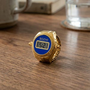 May include: A gold-toned digital ring watch with a blue face displaying the time 5:21. The watch has the brand name "BOZHI" above the digital display. The ring band is also gold-toned and appears to be adjustable.