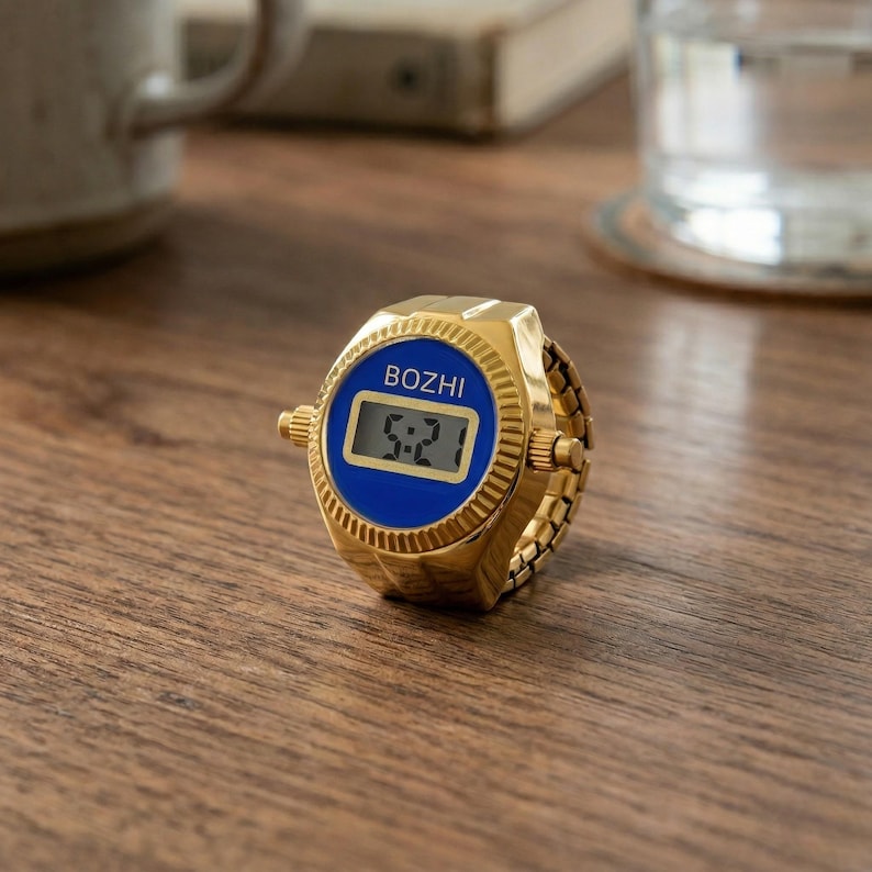 May include: A gold-toned digital ring watch with a blue face displaying the time 5:21. The watch has the brand name "BOZHI" above the digital display. The ring band is also gold-toned and appears to be adjustable.