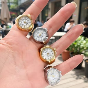 May include: Five ring watches are displayed on a hand, featuring white faces and gold or silver metal bands. The watch rings have an analogue design with numbers and hands. These rings are a unique fashion accessory.