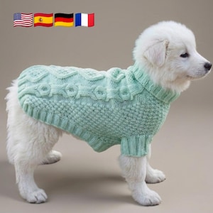 May include: A light mint green knitted dog sweater featuring a textured cable knit pattern along the back and a ribbed collar and hem. The garment is worn by a white fluffy puppy standing in profile against a solid neutral background.