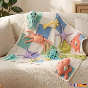 Puede incluir: Una manta tejida con un patrón de cuadros en blanco, azul claro y verde azulado. La manta está decorada con criaturas marinas tejidas en tres dimensiones, incluyendo un cangrejo color melocotón, dos estrellas de mar lavanda, dos pulpos azul claro, dos peces naranjas y vida marina verde.