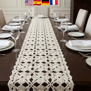 Puede incluir: Camino de mesa de ganchillo color crema con un patrón floral, colocado sobre una mesa de comedor marrón oscuro preparada para una comida. La mesa está puesta con platos, vasos y cubiertos blancos.