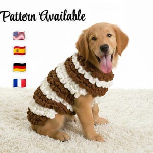 May include: A Golden Retriever puppy wearing a handmade brown and white crochet dog sweater. The sweater has a textured, layered design. Text at the top reads "Pattern Available" with flags of the USA, Spain, Germany, and France.