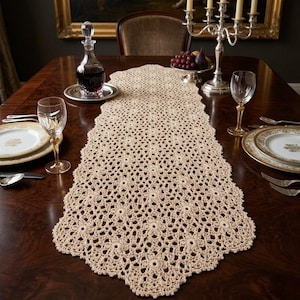 May include: A beige, crocheted table runner with a scalloped edge, displayed on a dark wood dining table. The runner is adorned with a delicate floral pattern. The table is set with fine china, silverware, and wine glasses, suggesting a formal dining setting.