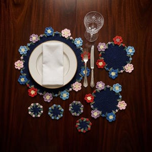 May include: A navy blue crochet placemat and coasters set with colourful flower accents. The placemat holds a white plate and napkin, with silverware and a water glass nearby. The coasters and placemat are decorated with red, blue, pink, and yellow flowers.