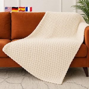 May include: A cream-colored throw blanket featuring a thick, chunky knit texture with a repeating basketweave pattern. The blanket is draped over a solid burnt orange sofa, showcasing the soft, raised yarn loops and the dense, woven construction.