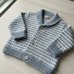 Peut inclure: Un cardigan pour bébé à motif quadrillé bleu clair et blanc. Le pull a un col châle, des poignets côtelés et trois boutons. L'étiquette sur le col indique "handmade".