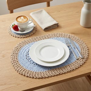 Peut inclure: Un set de table en crochet avec un centre bleu clair et une bordure beige, avec des assiettes blanches, une fourchette et un couteau. Un dessous de verre assorti contient une tasse de café et une fraise.