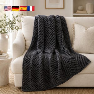May include: A dark charcoal grey knitted throw blanket featuring a prominent chevron or herringbone pattern with an open, airy stitch construction. The blanket is draped over a light-coloured sofa, showcasing its textured, thick yarn design.